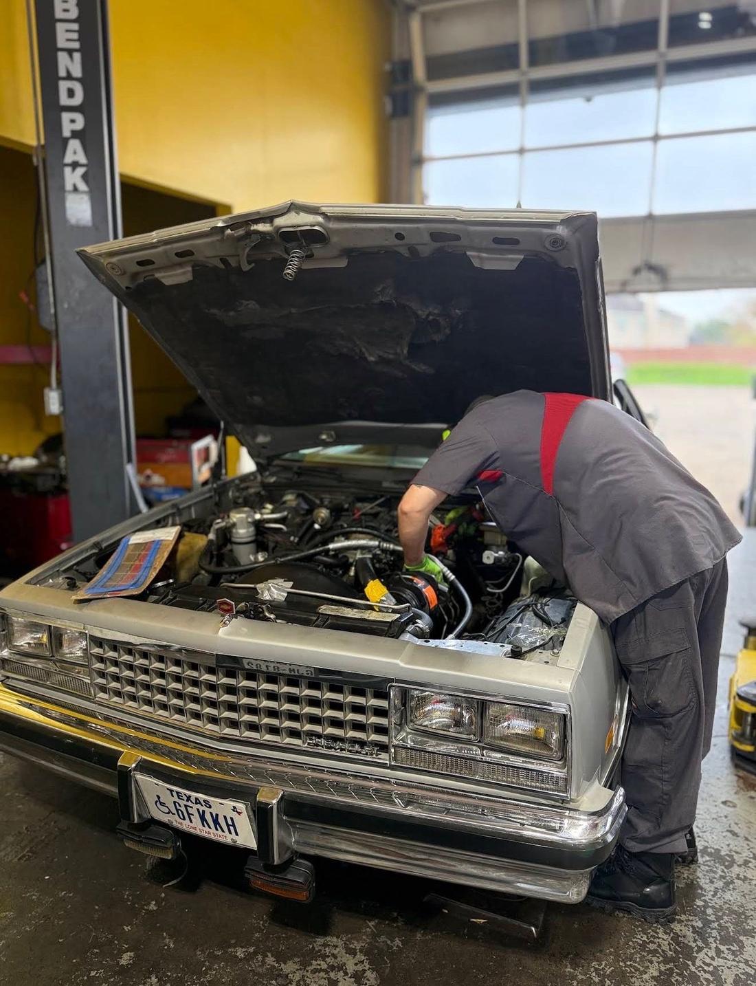 Technician leaning under the open hood of a silver classic Chevrolet El Camino inside the shop bay