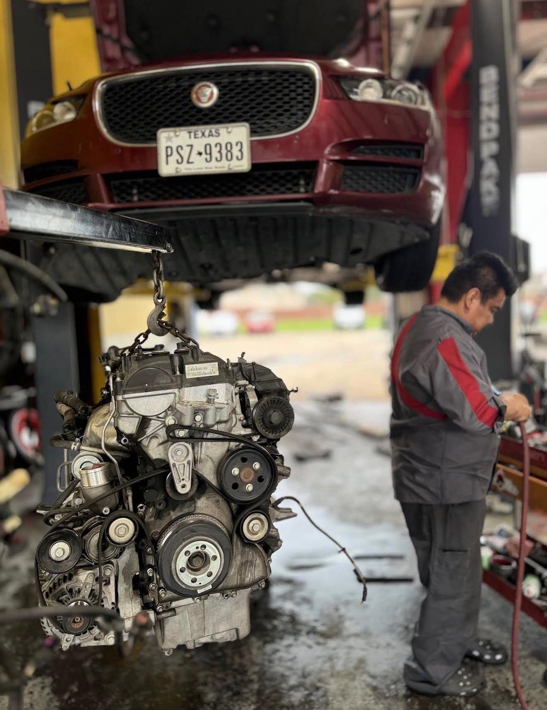 Engine hanging from a hoist chain below a burgundy Jaguar on the lift while a technician works