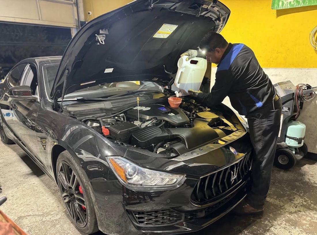 Technician wearing a headlamp pouring fluid under the open hood of a black Maserati